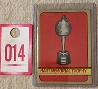 Front side close-up of the 1972-73 O-Pee-Chee Hart Memorial Trophy hockey card featuring the trophy image with red background and yellow banner labeled 'HART MEMORIAL TROPHY'. Card is in plastic protector and placed next to a tag for scale.
