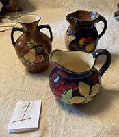 Three vintage pottery pieces: two pitchers and one handled vase, all hand-painted with fruit and floral designs on a beige textile surface.