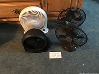 Four table top fans placed on carpet: one white Honeywell fan, one black round fan, and two black vintage-style metal blade fans.
