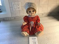 Front view of the vintage Pedigree doll wearing a red floral dress and lace bonnet sitting on a counter.