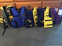 Photo of seven life jackets arranged in a row showing varying colors and designs to illustrate the lot contents.