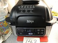 Front view of Ninja AG301C 4-in-1 indoor grill with digital control panel and Ninja logo on top lid, along with cooking tips label.