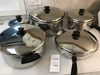 Set of five stainless steel pots and pans with lids arranged on a countertop showing their sizes and reflective surfaces.