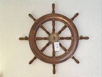 Clear front view of the large wooden ship's wheel with 8 spokes and central brass hub hung on a plain wall.