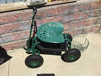 Green tractor-style plastic work seat on a metal frame with four large pneumatic tires. Front wheels are steerable. It has a black plastic tool tray below the seat and a metal mesh basket mounted on the back. The frame includes a retractable handle for pulling the seat along.