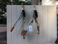 Two come along hand winches hanging on a white wooden fence outdoor, showing full side views of each tool with metal bodies, black ratchet housing, hooks, and different colored handle grips (orange and red).
