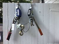 Two come along hand winches hanging on a white fence, one with blue housing and one with black housing and orange handle