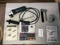 Photo of Bosch 4 1/2" corded angle grinder, side auxiliary handle, lock-on slide switch, two wrenches, protective guard, and manuals arranged on bubble wrap.