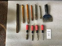 Photo of 11 metal chisels in various sizes and types laid out on a white cloth surface, showing different handles and blade widths.