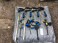 Eight clamps of various sizes and handle colors laid out on a grey tarp on gravel