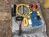 Photo of two grease guns, two ropes, one hatchet with head cover, six tie down straps of different sizes, and a 16-foot extension cord arranged on a gray tarp.