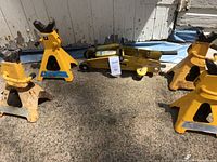 Photo shows one yellow hydraulic floor jack and four yellow 3-ton jack stands arranged on a concrete surface against a weathered wall.