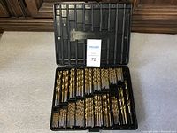 Open black plastic case showing a large collection of gold-colored drill bits arranged by size in individual slots.