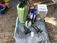 Four pressure sprayers of different sizes, four green metal hose guides, and Wood Shield Spray Applicator box shown arranged on a tarp outdoors