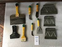 Ten assorted scrapers with metal blades and yellow handles with green tips arranged on a white towel for display.