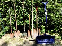 Seven garden tools including shovels, rakes, hoes with yellow tape on handles and a large blue plastic shovel grouped against greenery outdoors.