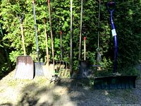 Nine garden tools arranged outside against greenery: various shovels, rakes, cultivator with red tape on handles, blue snow shovel visible.