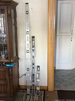 Three aluminum spirit levels of varying lengths (6', 4', 2') leaning against wall beside wooden cabinet