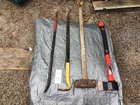 Photo showing four manual tools laid out on a tarp: axe, sledge hammer, pry bar, and crowbar.