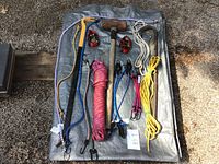 Top-down view of the laid out tools and hardware on a tarp showing sledgehammer, crowbar, several pry bars, nylon ropes coiled in pink, white, and yellow, four elastic bungee cords with hooks, and two tie down straps.