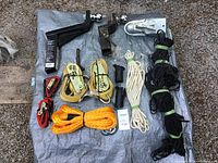 Photo of the full lot showing two trailer hitches with attached balls, ratchet tie downs in yellow and red, white and black ropes, thick orange tow rope, black cargo net, and black hitch cover organized on a tarp on gravel ground.