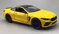 Yellow BMW M8 Competition diecast model car showing left side with black wheels and roof, front grille details, and side air vent.