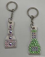 Photo of two white plastic lottery scratcher keychains attached to silver metal split rings and chains. One keychain decorated with pink, purple, and clear rhinestones; the other with green and purple rhinestones.
