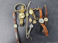 Top view of lot of 10 women's wristwatches arranged on black surface, showing variety of band materials and face styles.