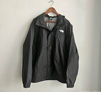 Front view of black rain jacket on hanger, showing elastic cuffs and front closure.