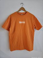 Orange Supreme T-shirt hanging on a wooden hanger against white background, featuring visible front logo.