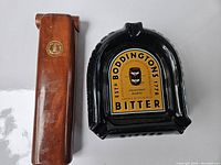 Solid wood case with gold emblem seen from top, alongside black ceramic ashtray with colorful Boddington's Bitter label.