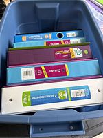 Large plastic bin containing assorted colorful binders stacked inside showing labels and brand stickers