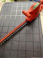 Full view of orange plug-in electric hedge trimmer placed on a black textured surface showcasing the blade and handle design.