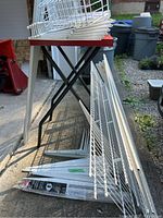 Stack of white metal wire shelving panels and various metal side brackets leaning against a table outdoors.