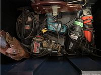 Photo showing assortment of power tools including angle grinder, jigsaw, drill, sander, and leather tool holster, cords attached, visible rust on some tools.
