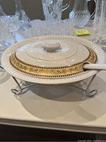 Oval porcelain casserole dish with lid, featuring 24k gold ornate trim, sitting on metal warming stand with ladle in place.