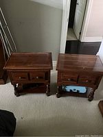 Two wooden side tables standing side by side on carpet showing front and side angles. Both have rectangular tops, two drawers each with round knobs, and stretcher shelves with carved legs.