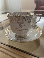 Side view of the cup showing the 50th Anniversary gold script and detailed gold floral pattern on white porcelain.