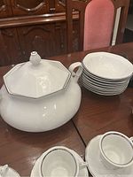 Photo showing 9 white soup bowls stacked next to a large white octagonal covered soup tureen on a wooden table.
