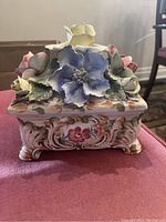 Front and side views of a rectangular ceramic jewelry box with floral decoration and raised ceramic flowers on the lid, showing detailed painting and some wear.