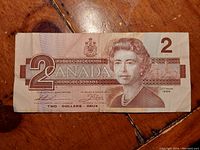 Front side of a 1986 Canadian two dollar bill showing Queen Elizabeth II portrait, denomination, and signatures, placed on a wooden surface.