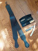 Full view of the antique shaving equipment including leather razor strap and two straight razors (one in pouch) on wooden floor