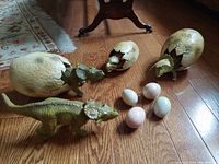 Two hatching dinosaur figures emerging from cracking eggs with three small stone eggs in front of them on wooden floor.