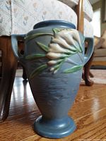 Front view of the vintage blue Roseville Freesia vase with raised white floral decoration and two handles, placed on wood floor.