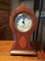 Front view of the wooden tabletop clock showing the clock face with black Arabic numerals and inlaid wooden floral design below.