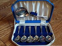 Set of six silver plated soup spoons with server displayed in case with satin and blue velvet interior.