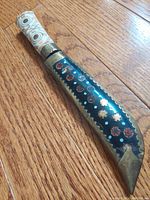 Dagger sheath showing the brass clad and colorful etched circular patterns with decorated handle in white with designs.