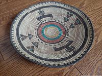 Top-side view of the round woven tray showing the intricate multicolored geometric pattern and concentric circle center design on wooden floor background.
