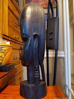 Side profile shot of the wooden bust showing detailed hair carving and ear with carved earrings, placed on a wooden surface against a brown cabinet background.