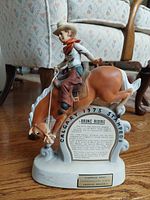 Front view of the ceramic decanter showing cowboy riding a bronco with detailed sculpting and painted finish. Base text reads 'Calgary 1975 Stampede'.